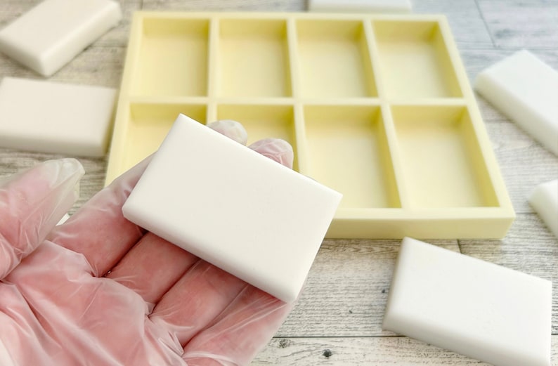 May include: A white rectangular silicone mold with six individual compartments for making soap bars. The mold is on a wooden surface, and a white soap bar is being held in a gloved hand.