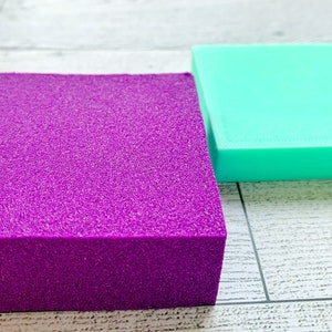 May include: Two rectangular blocks of foam, one purple and one turquoise, are sitting on a white wooden surface.