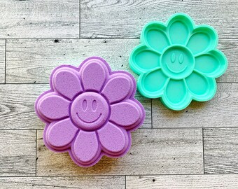 Op de afbeelding: Twee bloemvormige mallen met smiley-gezichten. Eén is lichtpaars, de andere is helder turquoise. De mallen zijn gemaakt van een flexibel materiaal, waarschijnlijk voor knutselen of bakken. De achtergrond is een lichte houtnerf.