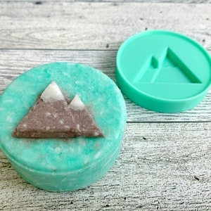 May include: A teal green round soap bar with a brown mountain design. The soap bar is next to a teal green silicone mold with a mountain design.