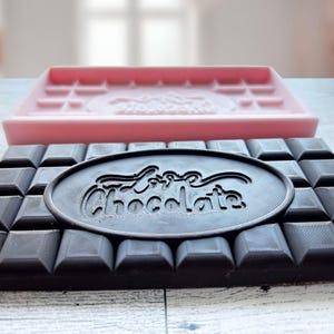 May include: A black chocolate bar mold with the words "Love Chocolate" embossed in a circle in the center. The mold is sitting on top of a pink silicone chocolate bar mold.