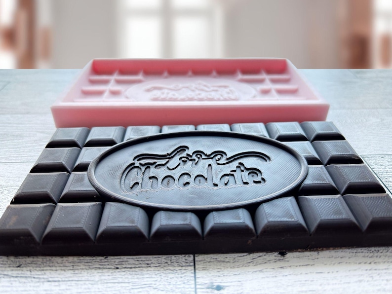 May include: A black chocolate bar mold with the words "Love Chocolate" embossed in a circle in the center. The mold is sitting on top of a pink silicone chocolate bar mold.