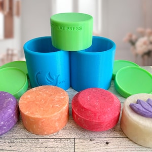 May include: Four round bars of soap in different colors, purple, orange, red, and white, with a flower design on the white bar. The soap bars are sitting on a wooden surface next to two green lids and two blue containers with the text "FLAT PRESS" on the top green lid.