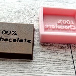 May include: Two chocolate bar moulds, one brown and one pink. The brown mould has the words "100% Chocolate" engraved on it. The pink mould has the words "100% Chocolate" engraved on it in reverse.