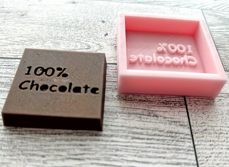 May include: Two chocolate bar moulds, one brown and one pink. The brown mould has the words "100% Chocolate" engraved on it. The pink mould has the words "100% Chocolate" engraved on it in reverse.