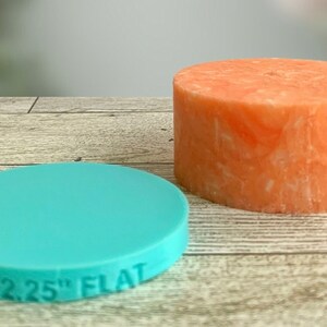 Pode incluir: Um molde redondo plano de silicone com 2,25 polegadas de diâmetro em azul turquesa, ao lado de uma vela redonda laranja com textura marmorizada.