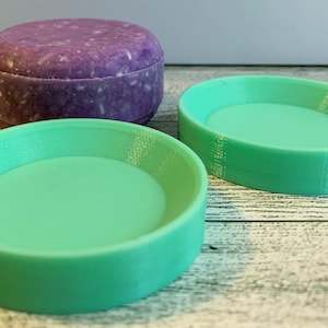 May include: A purple, round shampoo bar sits next to two mint green soap dishes. The shampoo bar has a speckled appearance. The soap dishes are round with a shallow basin and a raised edge. The items are on a light wood-grain surface.