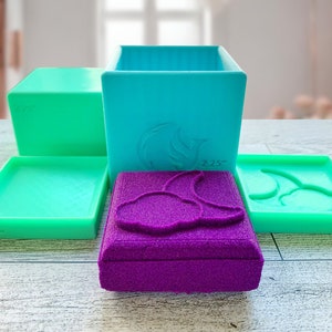 May include: Four silicone molds for making soap or resin crafts. The molds are in various shapes, including a square, a rectangle, and two different shapes with a curved design. The molds are green and purple.
