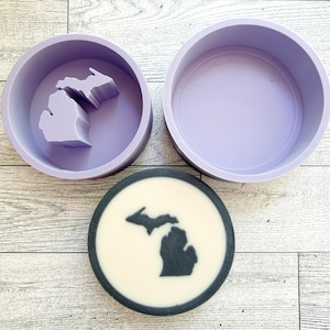May include: A purple silicone mold shaped like the state of Michigan. The mold is empty and ready to be used to make a soap or candle. A finished soap or candle is shown in the background, with a black outline of the state of Michigan on a white background.