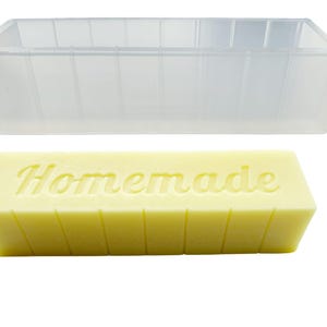 May include: A clear rectangular soap mould is above a yellow bar of soap. The soap has the word "Homemade" embossed on it. The mould is empty and has vertical lines on the sides.