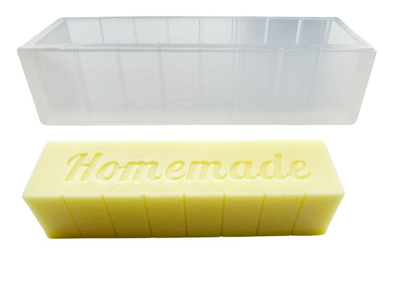 May include: A clear rectangular soap mould is above a yellow bar of soap. The soap has the word "Homemade" embossed on it. The mould is empty and has vertical lines on the sides.