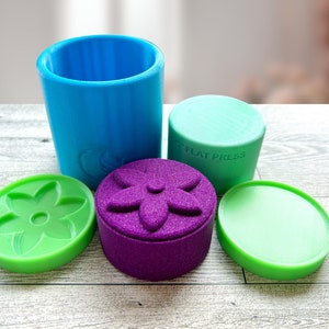 May include: A set of four silicone molds for making soap. The molds are in the shapes of a flower, a cylinder, and a circle. The molds are green, purple, and blue.