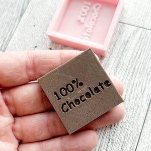 May include: A brown square silicone mould with the text "100% Chocolate" embossed in the centre. The mould is sitting on a wooden surface.