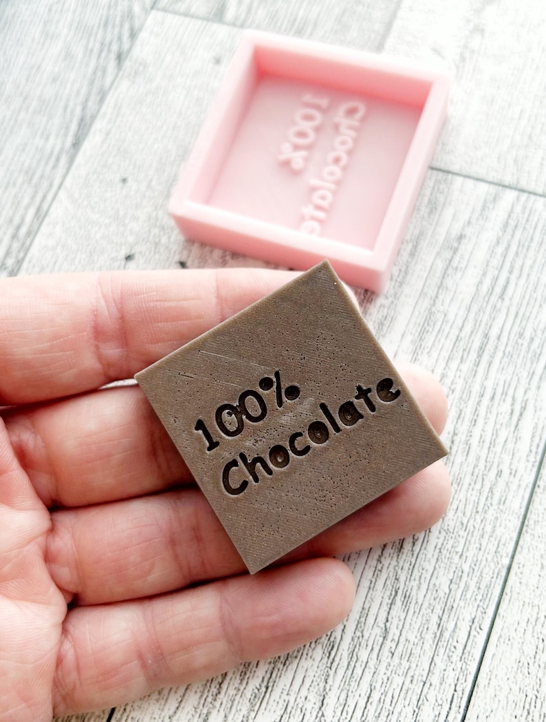 May include: A brown square silicone mould with the text "100% Chocolate" embossed in the centre. The mould is sitting on a wooden surface.