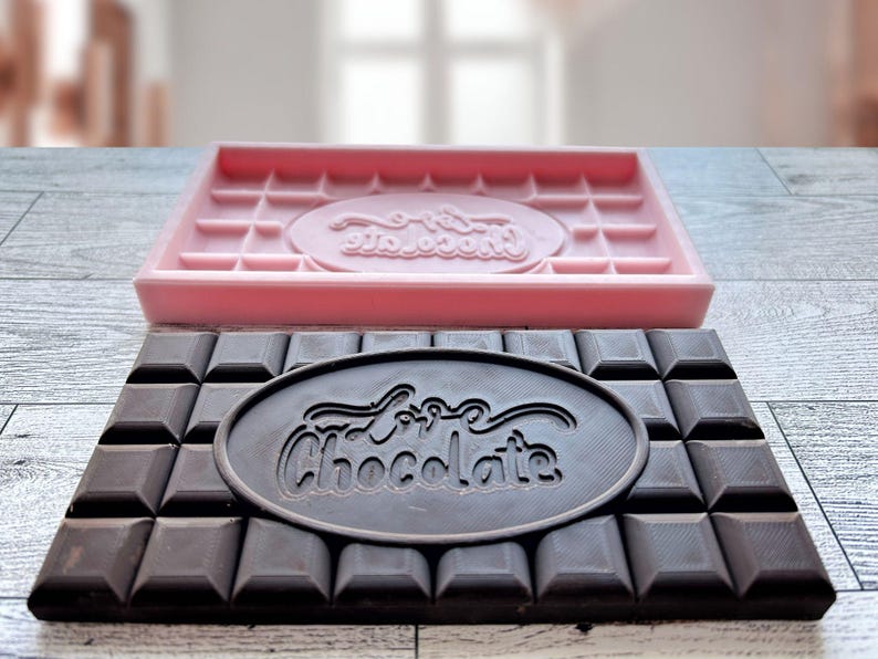 May include: A pink silicone chocolate mold with the words "Love Chocolate" embossed in the center. Next to it is a dark chocolate bar with the same words embossed in the center.