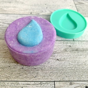 May include: A purple and blue soap bar with a blue teardrop shape on top. The soap bar is in a teal silicone mold.