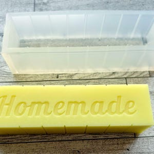 May include: A clear rectangular soap mould is positioned above a yellow soap bar with the word "Homemade" embossed on it. The mould is made of a translucent material and has a rectangular shape with vertical lines. The soap bar is a light yellow colour.