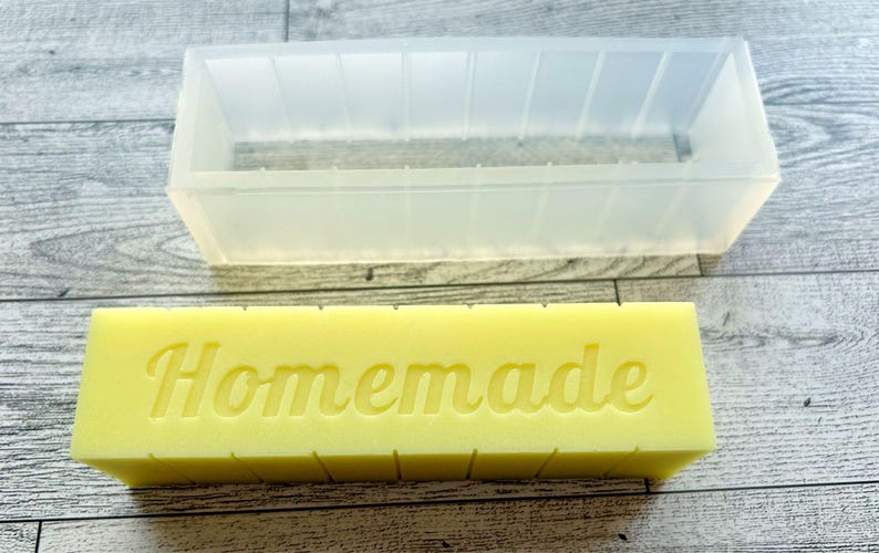 May include: A clear rectangular soap mould is positioned above a yellow soap bar with the word "Homemade" embossed on it. The mould is made of a translucent material and has a rectangular shape with vertical lines. The soap bar is a light yellow colour.
