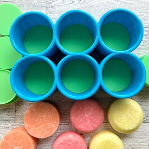 Pu&ograve; includere: Vista dall'alto di sei contenitori cilindrici blu con interni verdi, disposti in due file da tre. Coperchi circolari verdi e saponette rotonde colorate in tonalit&agrave; arancioni, rosa e gialle sono sparsi intorno ai contenitori.