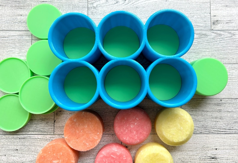 Pu&ograve; includere: Vista dall'alto di sei contenitori cilindrici blu con interni verdi, disposti in due file da tre. Coperchi circolari verdi e saponette rotonde colorate in tonalit&agrave; arancioni, rosa e gialle sono sparsi intorno ai contenitori.