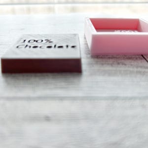 May include: A pink rectangular box with a lid, the lid is open and the box is empty.  The box is sitting on a white wooden surface.  Next to the box is a dark brown rectangular block with the words "100% Chocolate" engraved on the top.