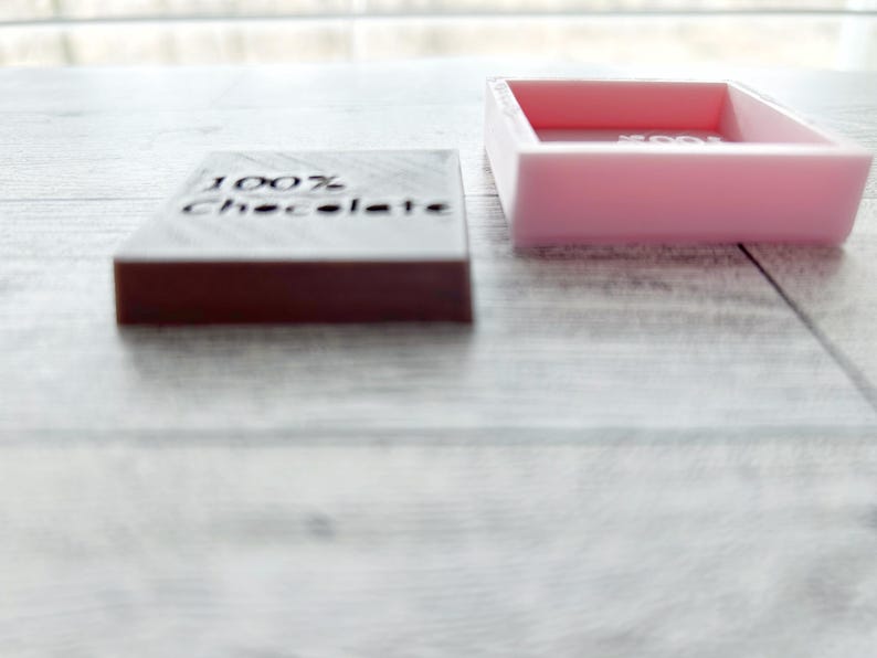 May include: A pink rectangular box with a lid, the lid is open and the box is empty.  The box is sitting on a white wooden surface.  Next to the box is a dark brown rectangular block with the words "100% Chocolate" engraved on the top.