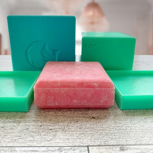 May include: Four teal green rectangular soap dishes, two of which are stacked, with a pink rectangular bar of soap in the center.