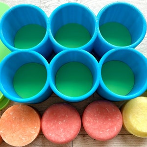 Pu&ograve; includere: Una vista dall'alto di una collezione di prodotti da bagno colorati. Ci sono sei supporti cilindrici blu con basi verdi, circondati da coperchi verdi e varie saponette per shampoo colorate nelle tonalit&agrave; dell'arancione, del rosa e del giallo. Gli articoli sono disposti su una superficie di legno chiaro.