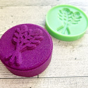 May include: Two round, purple and green bath bombs with a floral design. The bath bombs are on a white wooden surface.