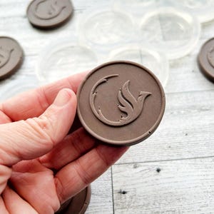 Peut inclure: Une main tient un objet circulaire marron avec un motif d'oiseau dans un croissant de lune. Plusieurs objets similaires et des moules transparents sont dispersés sur une surface claire à grain de bois. Les objets semblent être des moules ou des éléments décoratifs.