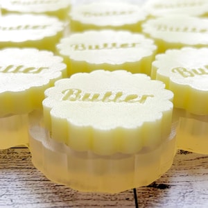 Può includere: Una collezione di saponette circolari giallo pallido con bordi smerlati disposte a griglia. Ogni singolo pezzo di sapone presenta la parola Butter impressa in un carattere chiaro e leggibile sulla superficie superiore. I saponi hanno una finitura opaca.