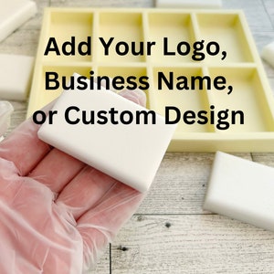 Puede incluir: Un molde de silicona rectangular blanco con múltiples compartimentos para hacer barras de jabón. El molde está sobre una superficie de madera. Una mano sostiene una barra de jabón blanca. El texto "Add Your Logo, Business Name, or Custom Design" es visible en la imagen.