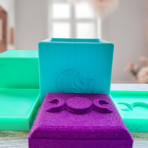 May include: A set of three teal blue and purple silicone molds for making resin crafts. The molds are square and rectangular with different designs, including a triple moon symbol and a stylized flame. The molds are 2.25 inches in size.