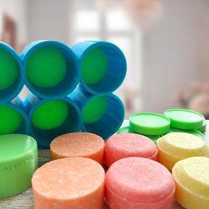 May include: A collection of colorful, round soap bars and cylindrical molds. The soaps are in shades of orange, pink, and yellow, with green and blue molds. One green soap has the text "2.25" FLAT PRESS".