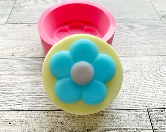 May include: A pink silicone mold for making a round soap or candle with a blue flower design. The mold is open and a yellow soap with a blue flower is sitting on top of it.