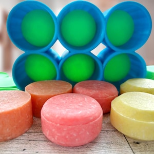 Pu&ograve; includere: Una collezione di saponette per shampoo rotonde e colorate, nei toni dell'arancione, del rosa e del giallo. Le saponette sono esposte con contenitori blu e verdi e coperchi verdi. L'immagine &egrave; scattata su una superficie di legno chiaro.