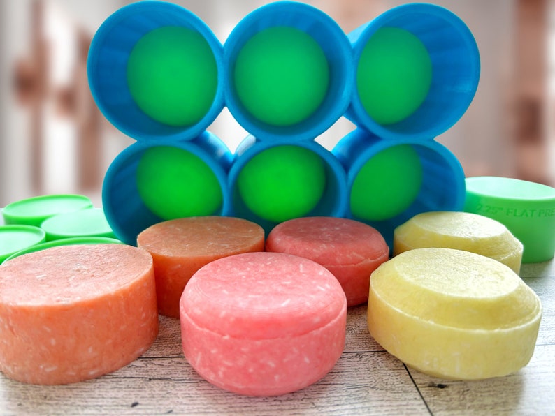 Pu&ograve; includere: Una collezione di saponette per shampoo rotonde e colorate, nei toni dell'arancione, del rosa e del giallo. Le saponette sono esposte con contenitori blu e verdi e coperchi verdi. L'immagine &egrave; scattata su una superficie di legno chiaro.