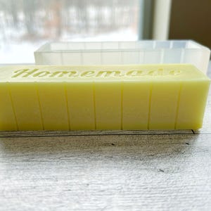 May include: A rectangular block of yellow soap with the word "Homemade" carved into the top. The soap is divided into sections with vertical lines. A clear, rectangular mould sits in the background.