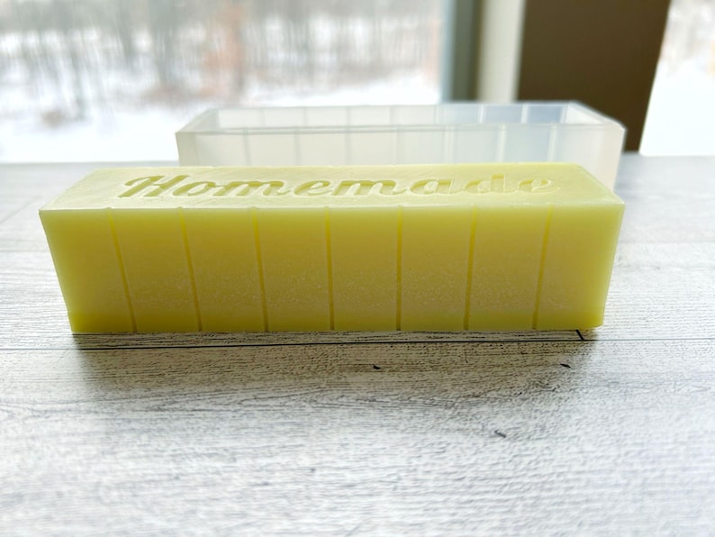 May include: A rectangular block of yellow soap with the word "Homemade" carved into the top. The soap is divided into sections with vertical lines. A clear, rectangular mould sits in the background.