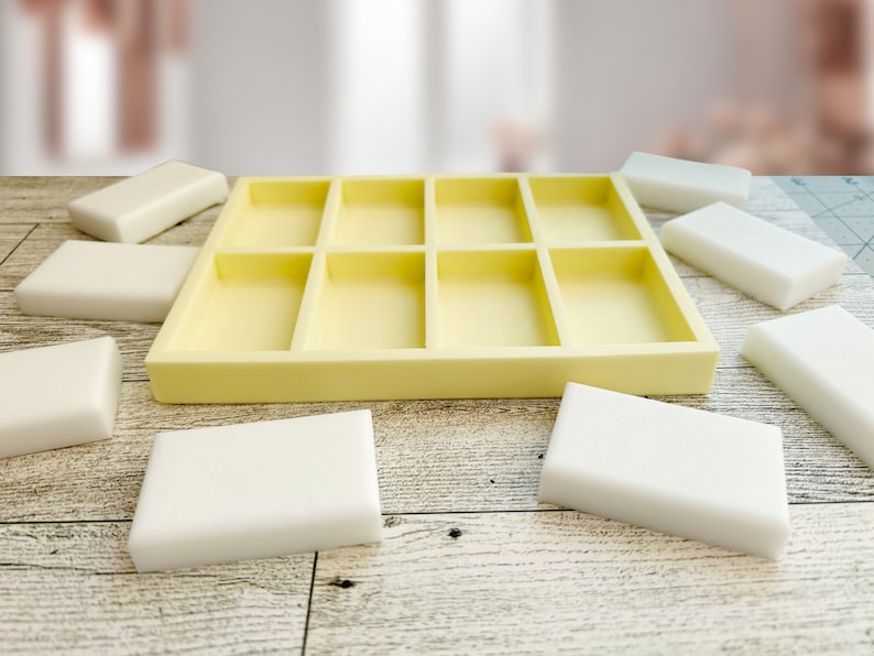 May include: A yellow silicone mold with nine rectangular compartments for making soap bars. The mold is sitting on a wooden surface with several white soap bars around it.