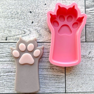 May include: A pink silicone mold of a cat paw with four toes and a paw pad. A gray cat paw with a pink paw pad is shown next to the mold.