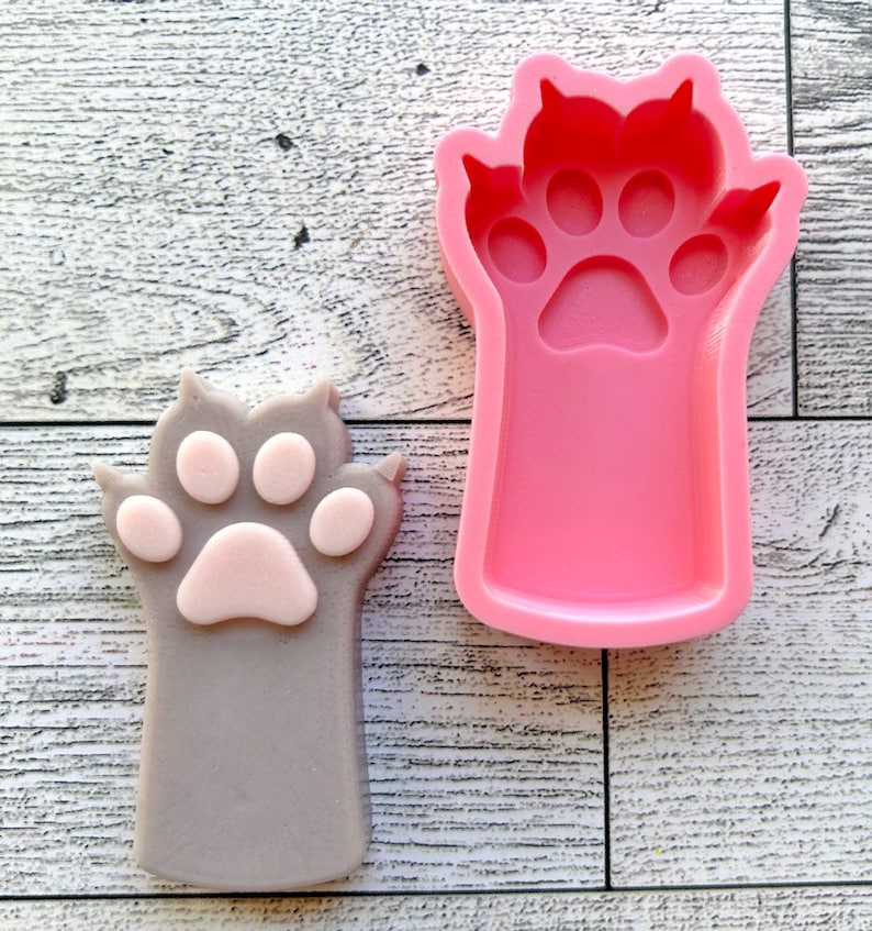 May include: A pink silicone mold of a cat paw with four toes and a paw pad. A gray cat paw with a pink paw pad is shown next to the mold.