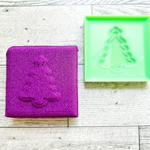 May include: A purple and green mold for making a Christmas tree shape. The mold is made of a textured material and has a raised design of a Christmas tree.