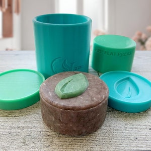 May include: A set of teal silicone molds for making soap. The molds are shaped like a round container, a flat circle, and a leaf. The molds are on a light wood surface.