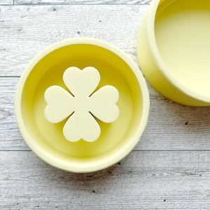 May include: A yellow ceramic dish with a four-leaf clover design. The dish is on a light wood surface.