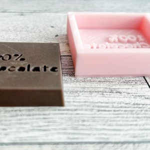 May include: A brown chocolate bar with the text "100% Chocolate" embossed on the top. Next to the chocolate bar is a pink silicone mould with the same text embossed on the inside.