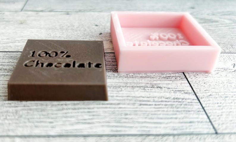 May include: A brown chocolate bar with the text "100% Chocolate" embossed on the top. Next to the chocolate bar is a pink silicone mould with the same text embossed on the inside.