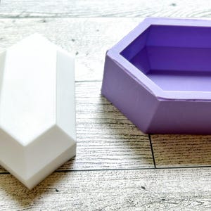 Puede incluir: Dos moldes de silicona para hacer formas geométricas. Un molde es un prisma hexagonal blanco y el otro es un prisma hexagonal morado.