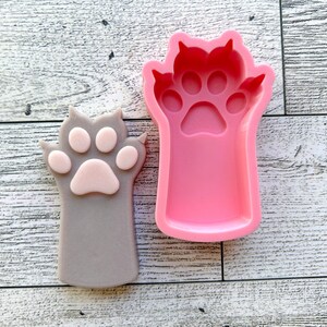May include: A pink silicone mold of a cat paw with four toes and a paw pad. Next to the mold is a gray cat paw with a pink paw pad.