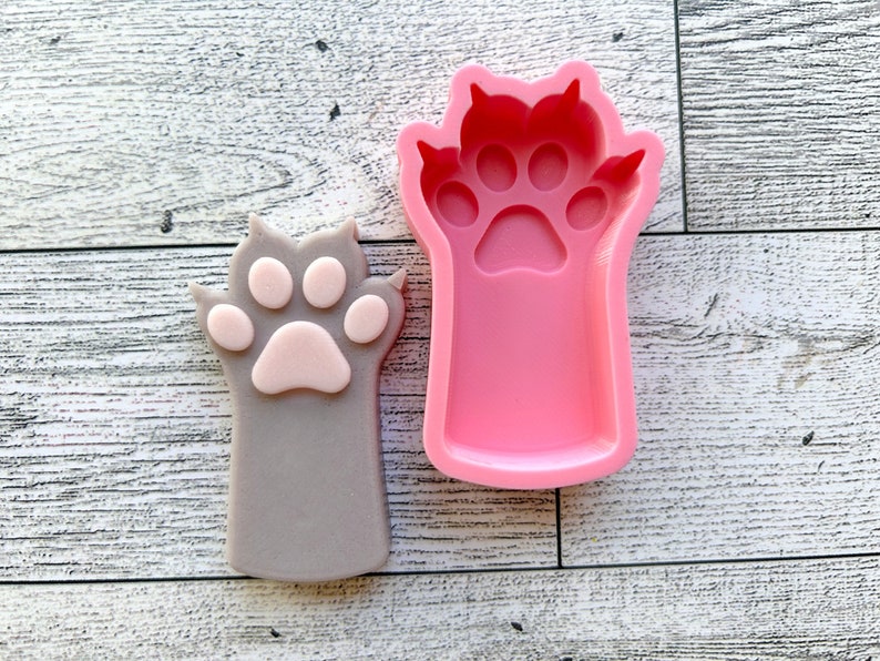 May include: A pink silicone mold of a cat paw with four toes and a paw pad. Next to the mold is a gray cat paw with a pink paw pad.